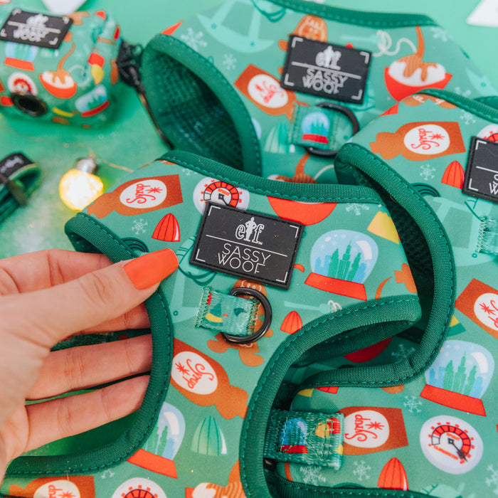A hand with orange painted nails holds the Sassy Woof Dog Adjustable Harness - Elf™, a green harness with festive holiday designs like snow globes, Christmas trees, and gifts. Similar Sassy Woof harnesses are visible in the background.