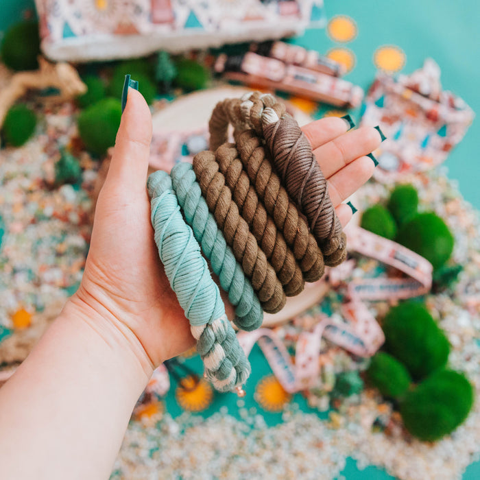 A hand holds several skeins of yarn in brown, tan, and teal—ideal for crafting the Dog Rope Leash - Tail Mix by Sassy Woof. The background shows beads, green moss, and ribbon rolls on a turquoise surface.