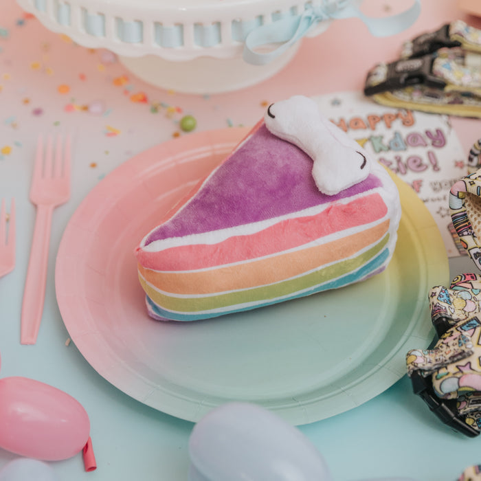 The Sassy Woof Dog Toy - Barkday Cake, a plush squeaky rainbow-layered cake slice with a white dollop on top, sits on a pastel plate surrounded by party decorations, balloons, and utensils for a festive dog celebration.
