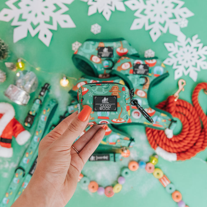 A hand with coral-orange nail polish holds a Sassy Woof Dog Waste Bag Holder - Elf™, featuring festive designs. Christmas-themed pet accessories, treats, and snowflake decorations are displayed on a green surface.