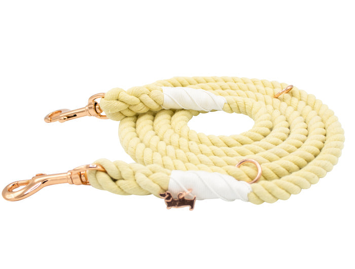 Introducing the Sassy Woof Hands-Free Rope Leash - Dandelion: a hand-crafted rope leash in a coiled, light yellow design, featuring rose gold metal clasps and a charming small dog-shaped charm. White tape securely fastens the ends of this adjustable leash, offering both style and durability.