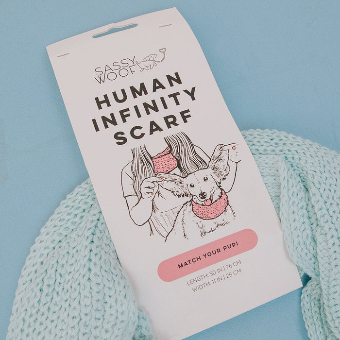 A product label shows a woman and her dog wearing matching scarves. The text reads: Sassy Woof Human Infinity Scarf - Blue. 50" x 11". A light blue knit dog scarf, perfect for cold weather, is displayed below the label.