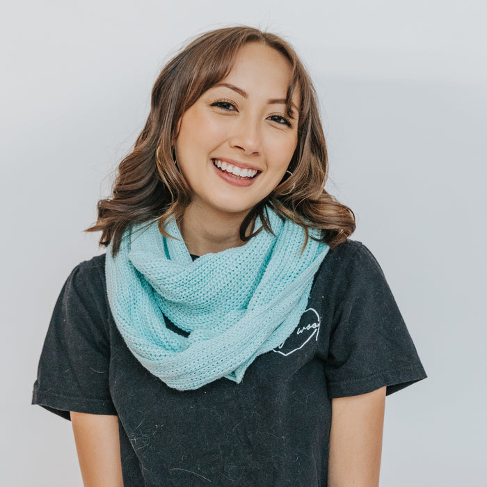 A young woman with wavy brown hair smiles at the camera, wearing a black t-shirt and the Sassy Woof Human Infinity Scarf in blue, standing against a plain white background.