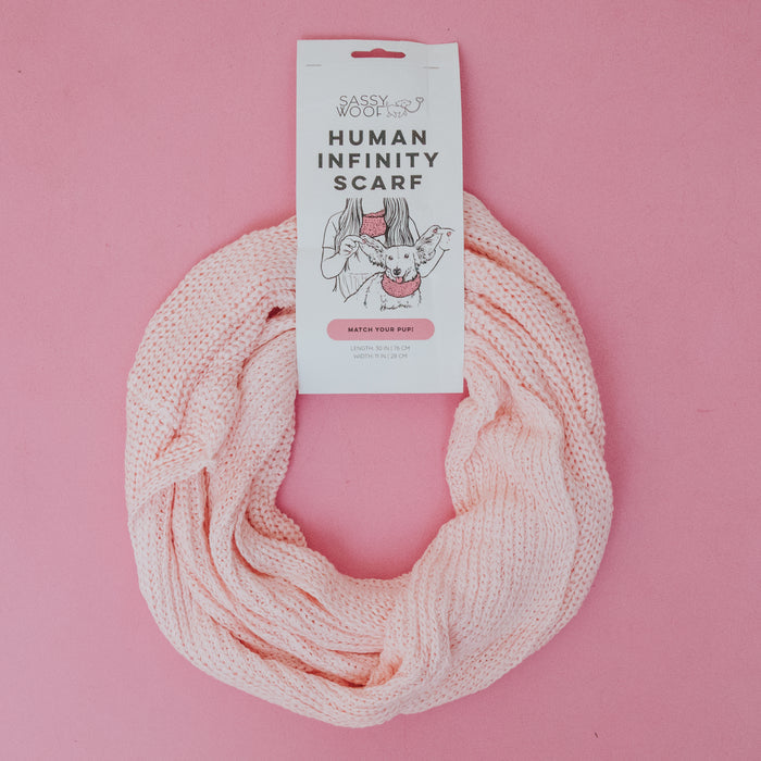 A light pink Sassy Woof Human Infinity Scarf is arranged in a circle on a pink background. Its label displays the brand and features an illustration of a person holding a dog—ideal for cold weather walks and coordinating with your pup.