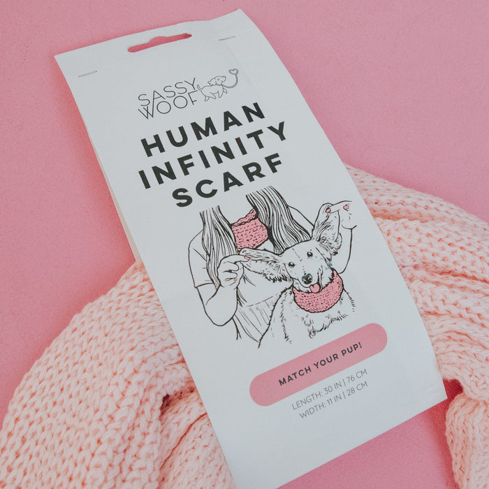 The Sassy Woof Human Infinity Scarf - Pink is pictured with its label showing a woman and her dog in matching scarves, displayed on a pink knitted scarf—perfect for chilly walks—against a pink background.