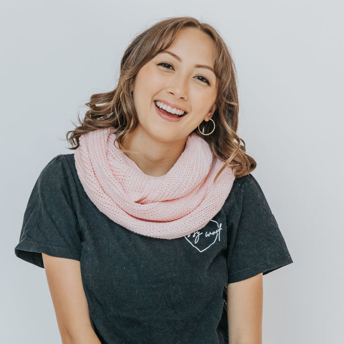 A smiling young woman with wavy brown hair models the Sassy Woof Human Infinity Scarf in pink, paired with a black t-shirt against a white background—ideal for chilly outdoor strolls.