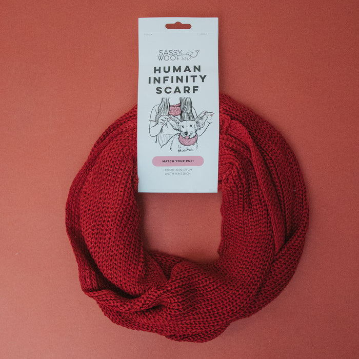 The Sassy Woof Human Infinity Scarf - Red is displayed in a circle on a red background with its branded tag, making it perfect for cozy cold weather walks with your pup.