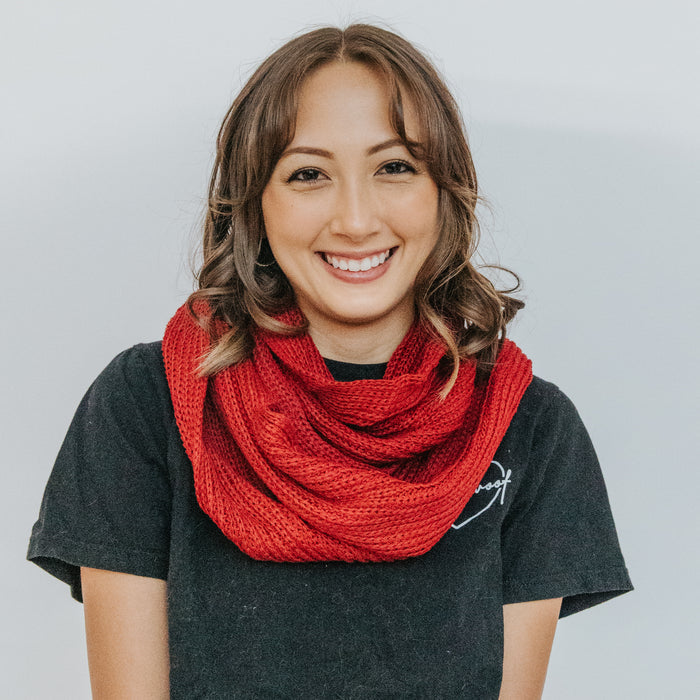 A woman with shoulder-length brown hair smiles at the camera, wearing a black t-shirt and the Sassy Woof Human Infinity Scarf in red—perfect for chilly days. She stands against a plain, light-colored background.