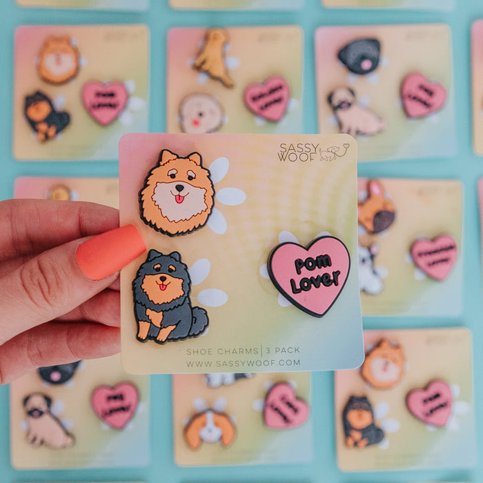 A hand with orange nail polish holds a Sassy Woof Shoe Charm - Poms card, featuring two cartoon Pomeranian charms and a pink heart reading "Pom Lover." Similar shoe charm cards are displayed on a blue surface in the background.